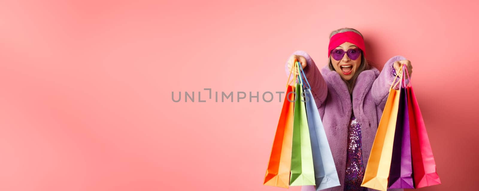 Happy asian adult woman going shopping, extend hands with shop bags and smiling cheerful, standing over pink background.