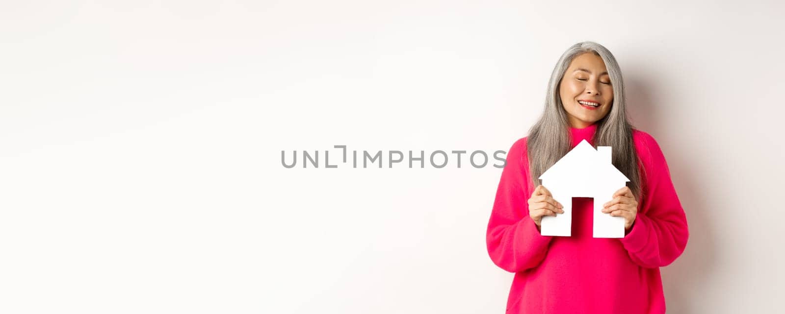 Real estate. Beautiful dreamy asian lady hugging paper house model with closed eyes, smiling as dreaming about buying apartment, standing over white background.