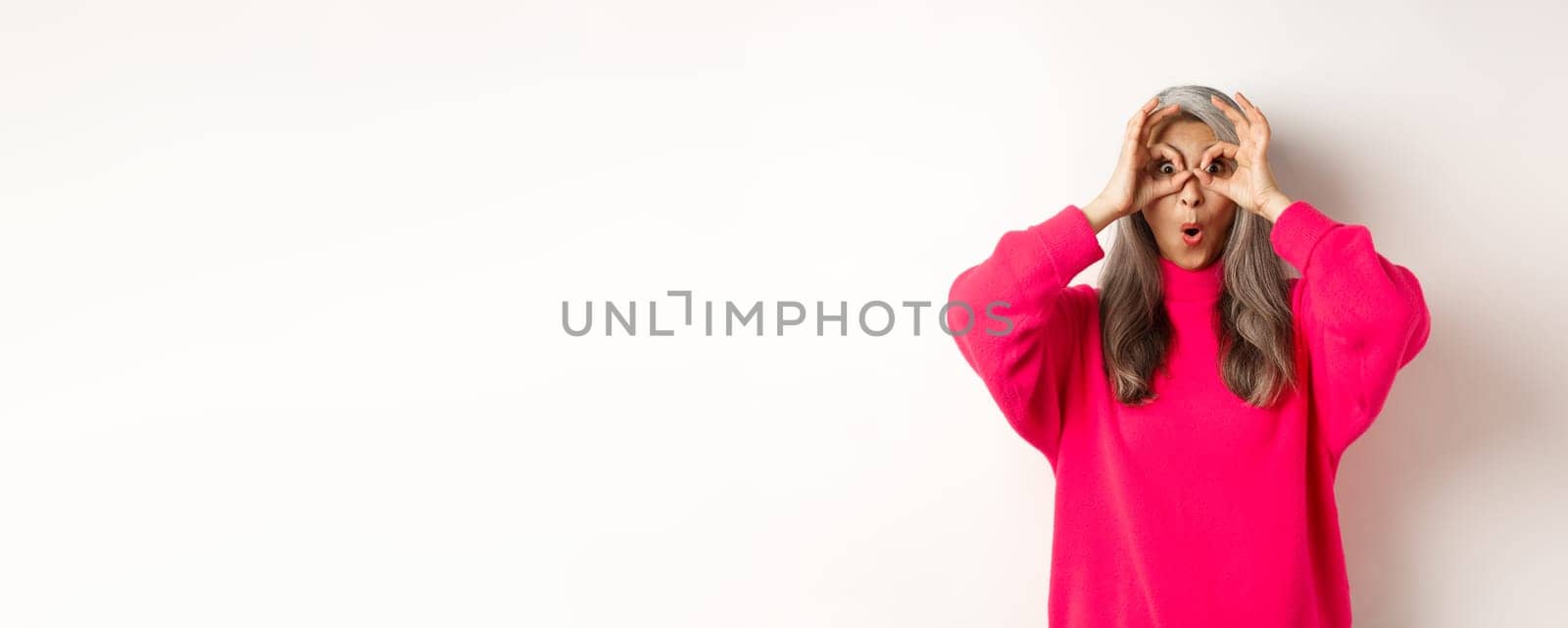 Image of funny asian grandmother looking through hand glasses with surprised and amazed face, checking out special offer, white background.
