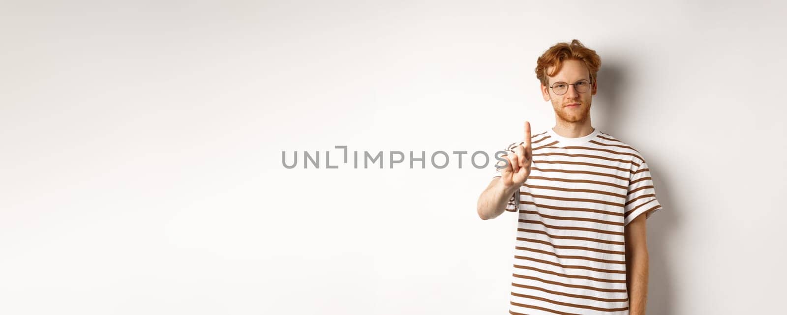 Young man with red hair and glasses shaking finger in prohibition, taboo gesture, disapprove and stop something, standing over white background.