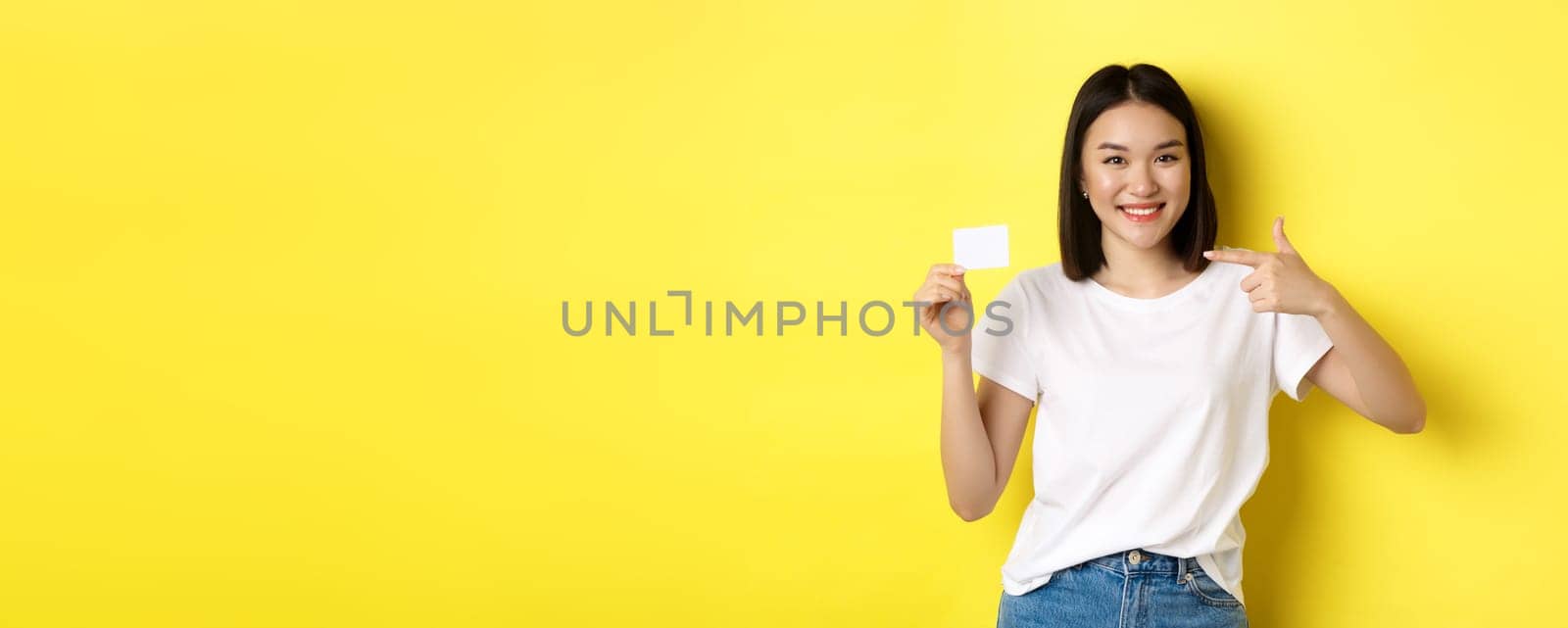 Young asian woman in casual white t-shirt showing plastic credit card and smiling at camera, yellow background.