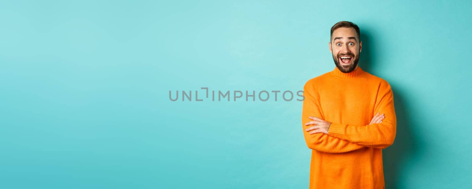 Image of happy and surprised man reacting to news, looking amazed, standing in orange sweater against turquoise background.