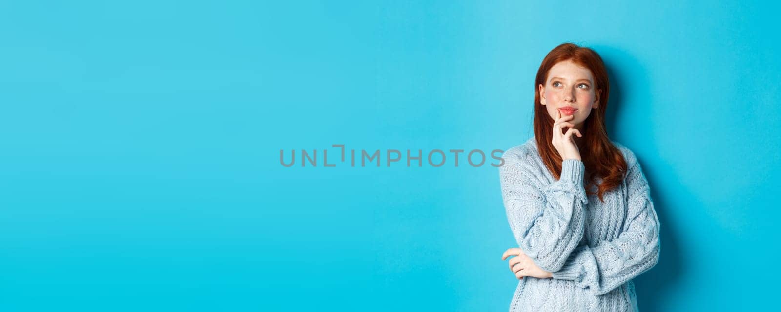 Thoughtful cute woman with red hair, looking upper left corner logo and thinking, imaging something, standing over blue background.