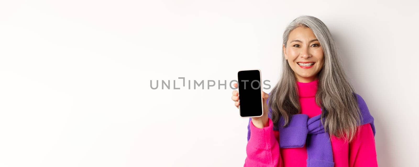 Online shopping. Close-up of smiling asian grandmother showing blank smartphone screen, recommending promotion, standing over white background.