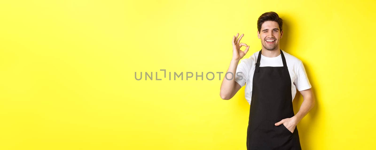 Confident and handsome waiter showing ok sign, wearing black apron and standing against yellow background.