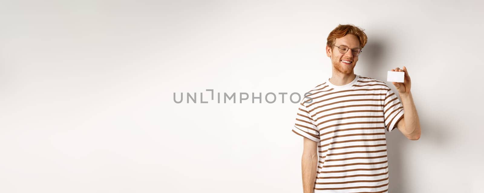 Shopping and finance concept. Cheerful redhead male student in glasses showing plastic credit card and looking satisfied, standing over white background.