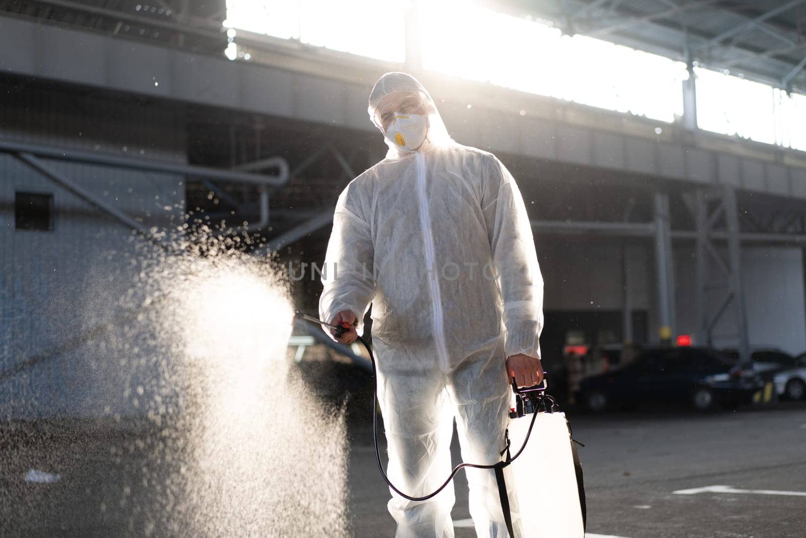 Man dressed white protective overalls spraying surface antibacterial sanitizer sprayer during quarantine Caucasian person protective suit disinfects parking during covid pandemic backlight