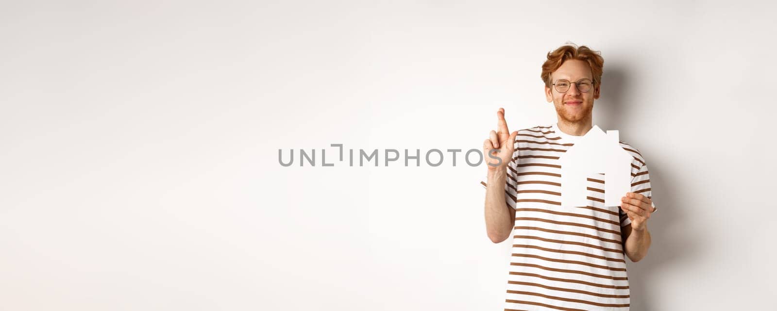 Hopeful redhead man dreaming of buying house, holding paper home cutout and cross fingers for good luck, making a wish, standing over white background.