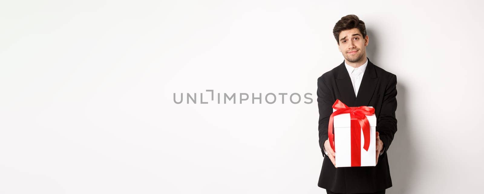 Concept of christmas holidays, celebration and lifestyle. Image of handsome boyfriend in black suit, giving you a present, extending hands with gift, standing over white background.