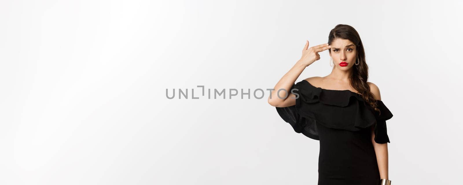 Fashion and beauty. Annoyed young woman in black dress making finger gun gesture near head, shooting herself from irritation, standing over white background.