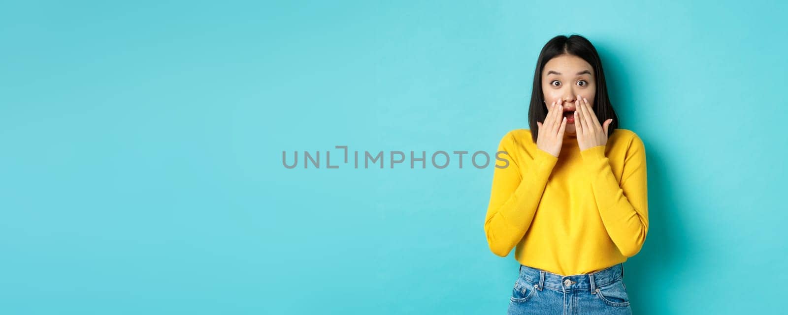 Portrait of impressed korean girl saying wow, gasping and staring amazed at camera, standing in yellow pullover over blue background.