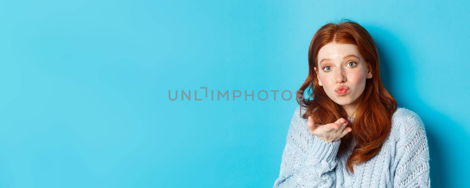 Silly redhead woman in sweater, blowing air kiss at camera with puckered lips, standing against blue background.