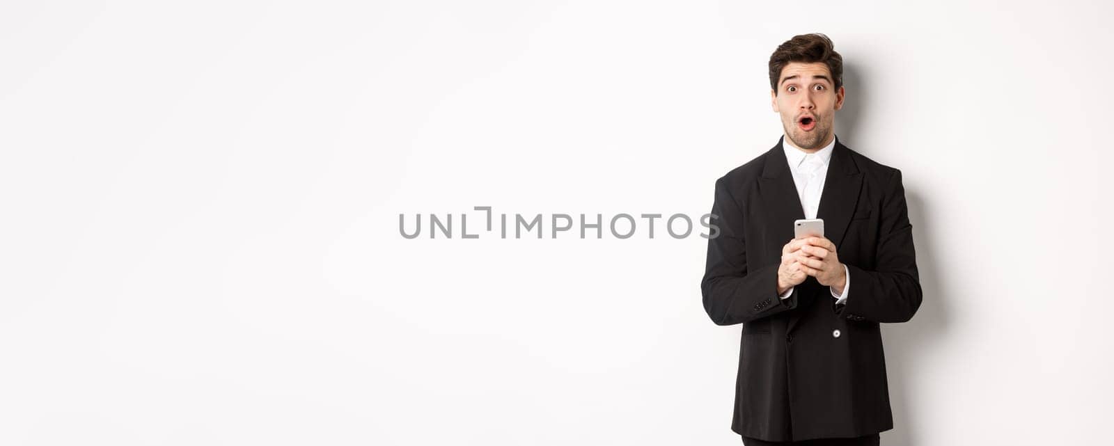 Amazed handsome guy in black suit reacting to cool promo offer, holding mobile phone, standing against white background.