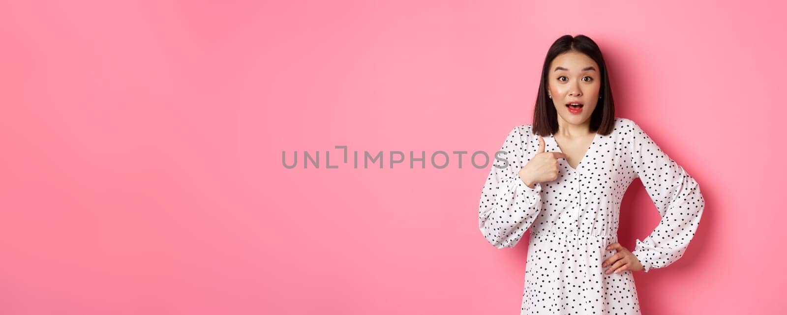 Beautiful brunette asian woman pointing finger at camera, looking surprised as being chosen, standing in dress against pink background by Benzoix