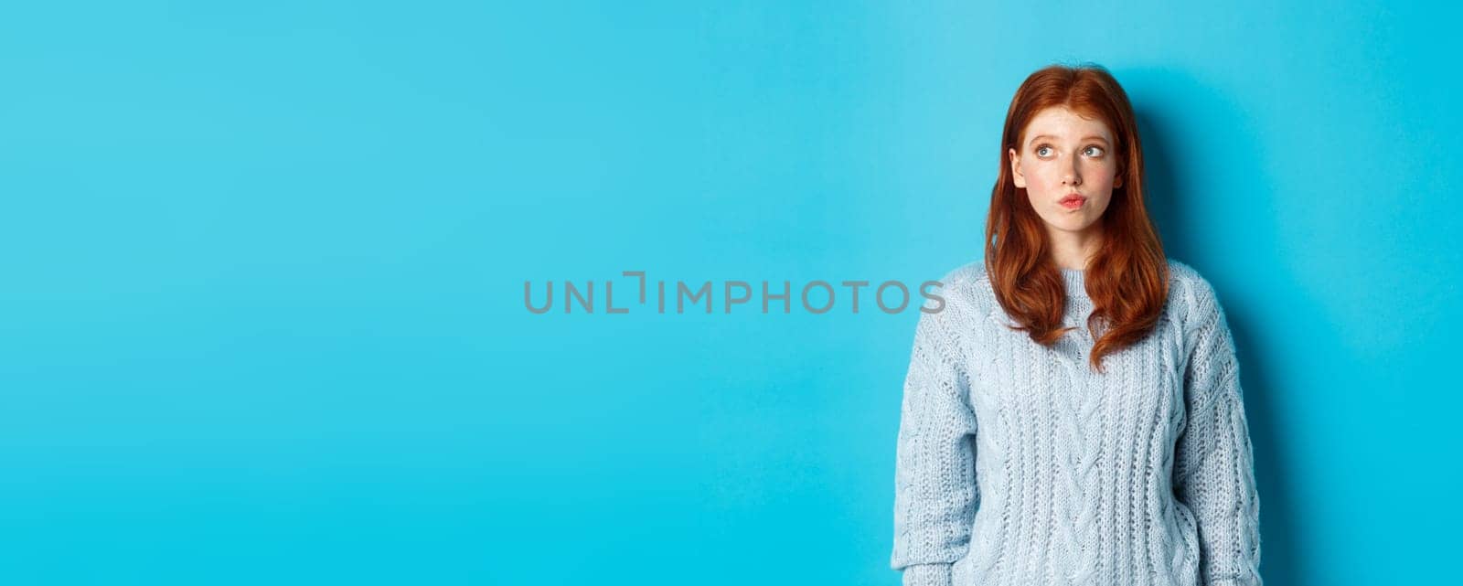 Dreamy redhead girl thinking or making decision, looking at upper left corner logo, standing against blue background.