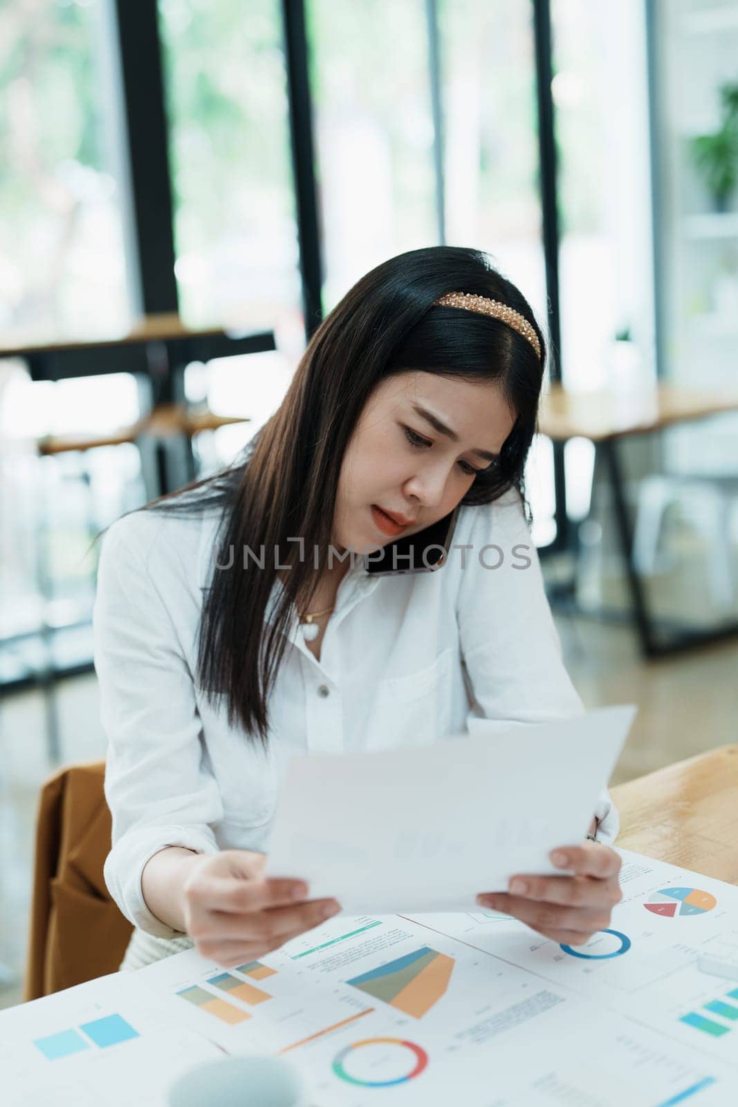 A portrait of a beautiful Asian female employee showing a stressed face while using the phone and financial documents on her desk.