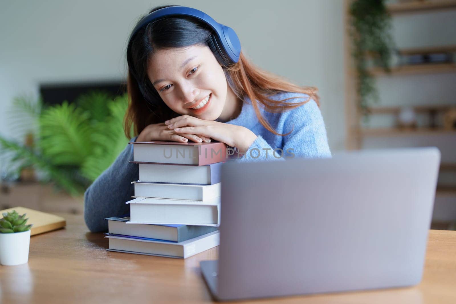 Portrait of young beautiful Asian woman showing smiling face during early morning online class with books, headphones and computer as study materials at home.