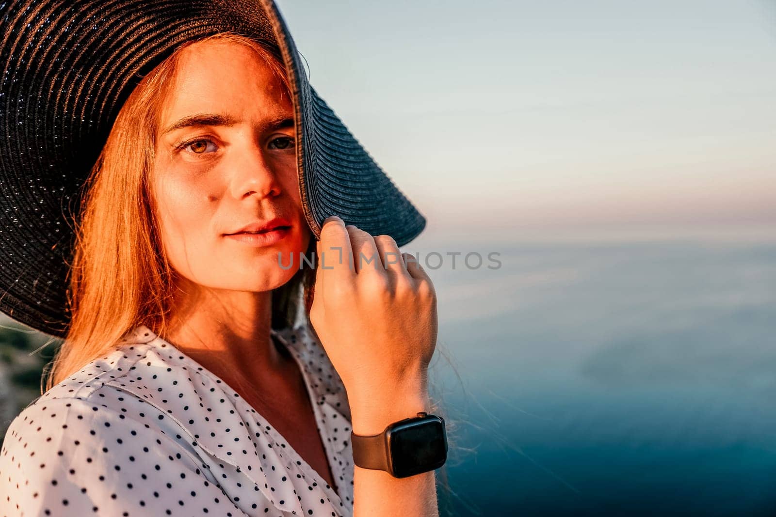 Portrait of happy young woman wearing summer black hat with large brim at beach on sunset. Closeup face of attractive girl with black straw hat. Happy young woman smiling and looking at camera at sea