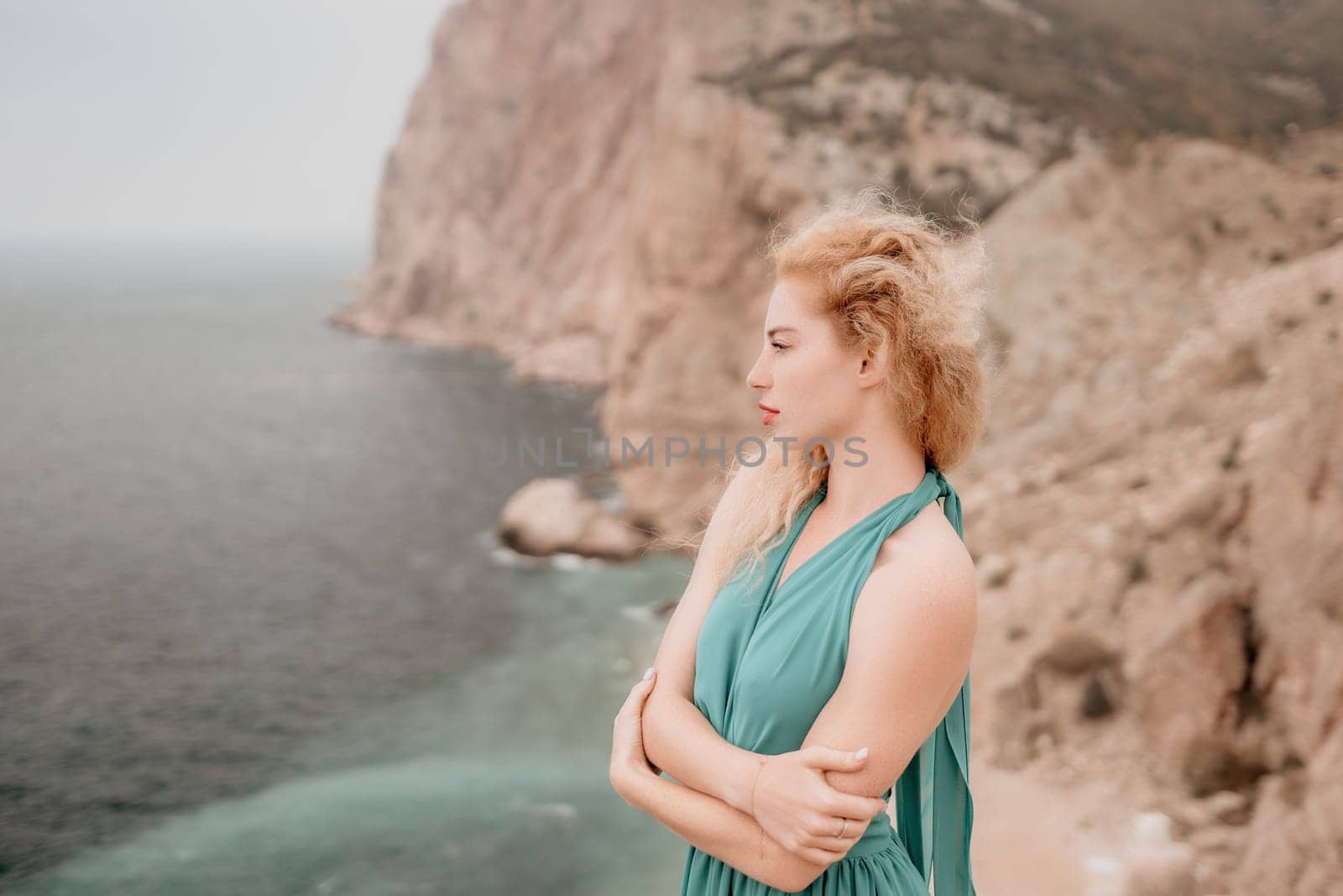 Side view a Young beautiful sensual woman in a mint long dress posing on a volcanic rock high above the sea during sunset. Girl on the nature on overcast sky background. Fashion photo