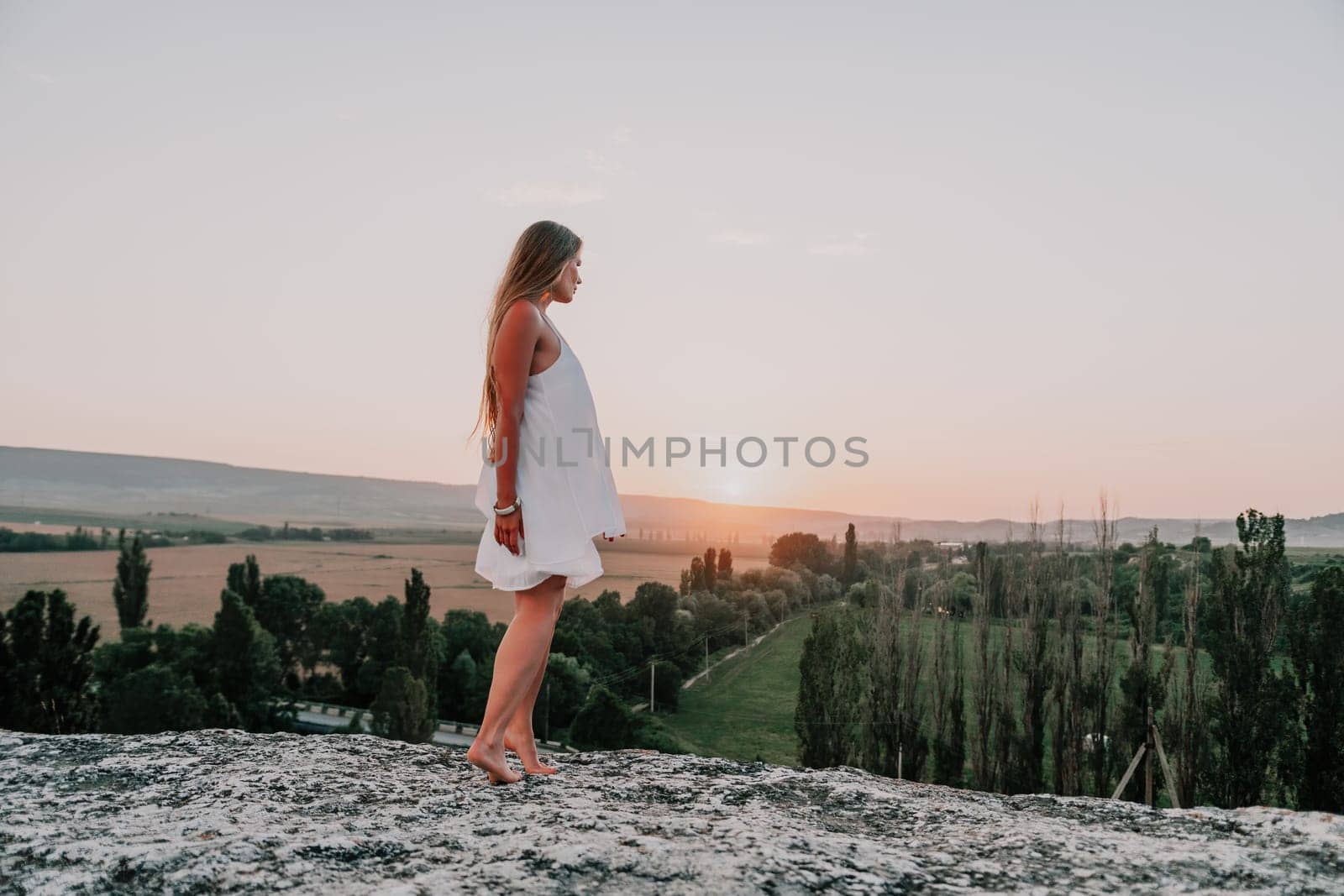 Happy woman standing with her back on the sunset in nature in summer with open hands. Romantic beautiful bride in white boho dress posing with mountains on sunset by panophotograph