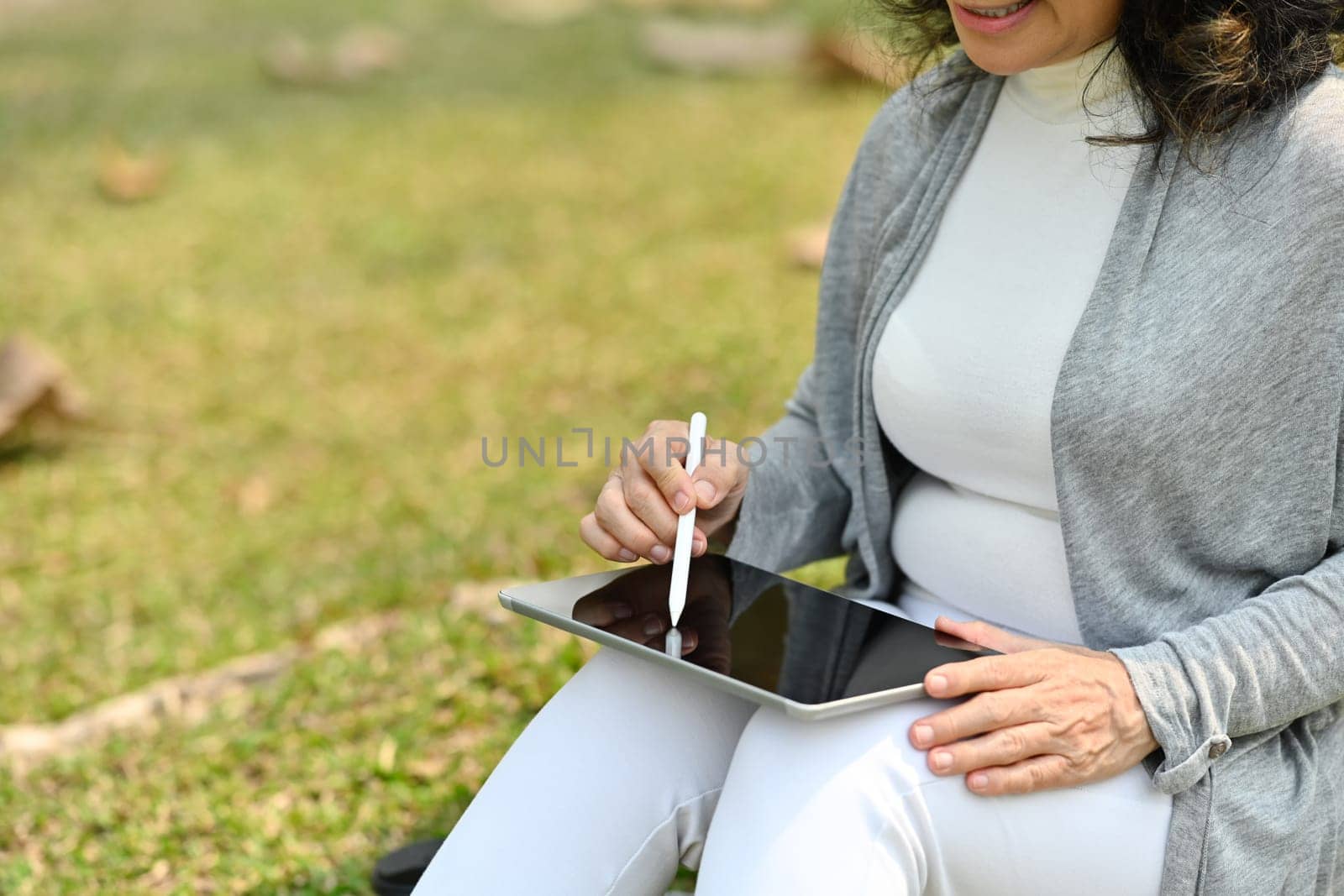 Senior woman using digital tablet on the grass outdoors in summer park. Retired lifestyle and technology concept.