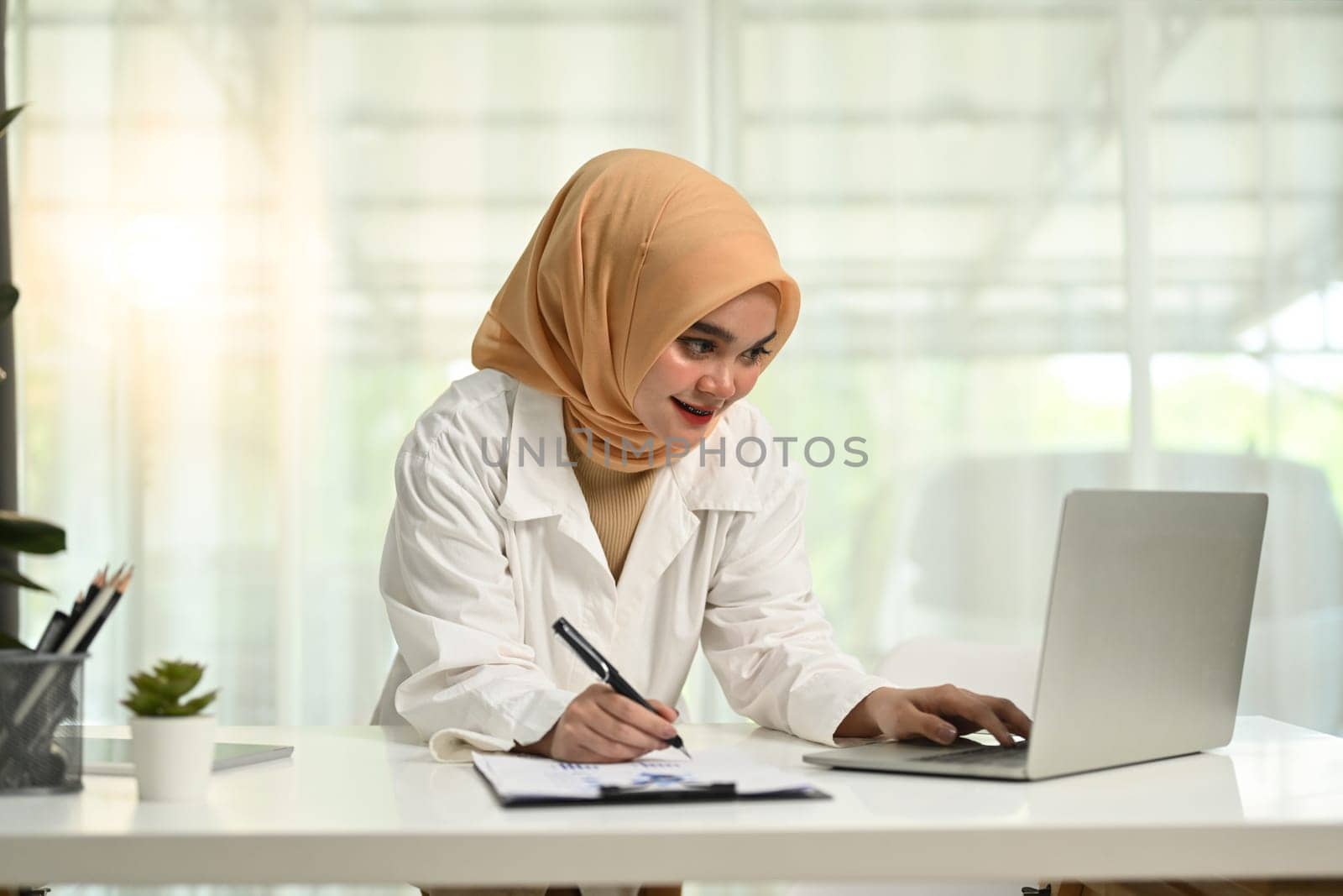 Portrait of smiling young muslim woman in hijab looking at laptop screen and writing information on document.