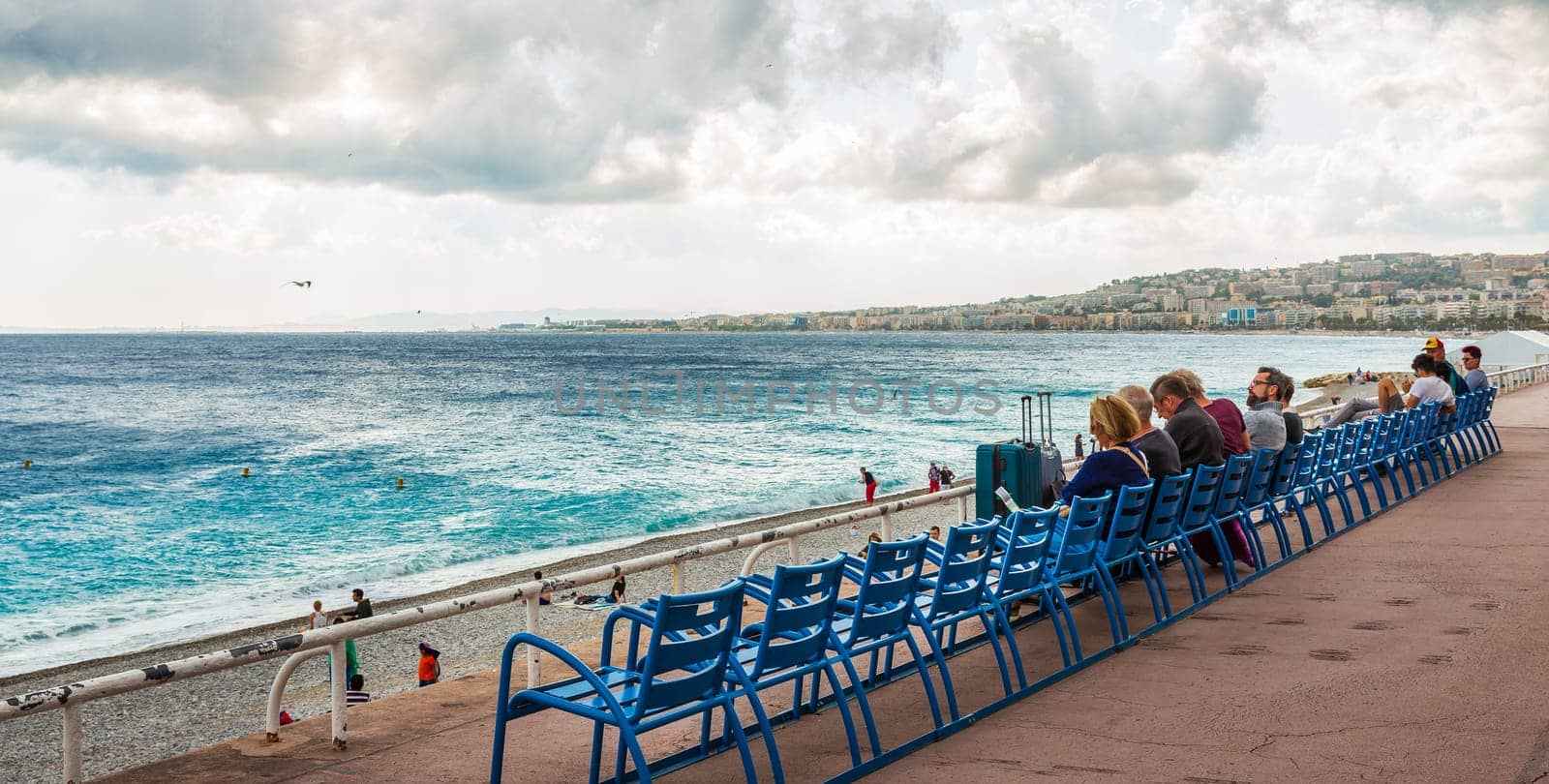 Nice, France - October 1, 2018: People are relaxing on blue seats on the beach of Mediterranean sea with background of city and cloudy sky.