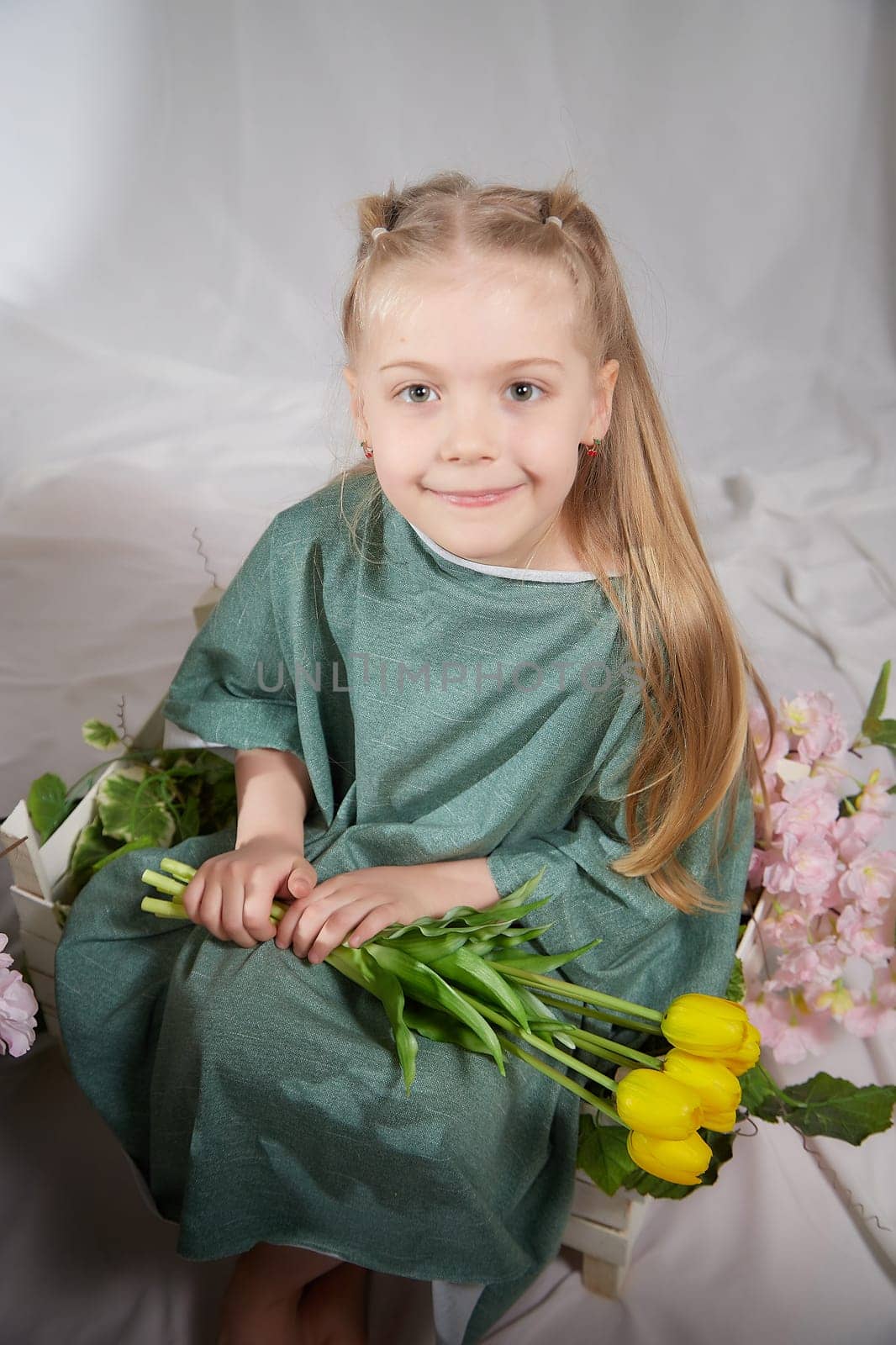 Portrait of a little blonde girl with bouquet of spring yellow flowers on a light background. Child in green dress holding a bouquet of tulips in hands. Spring concept