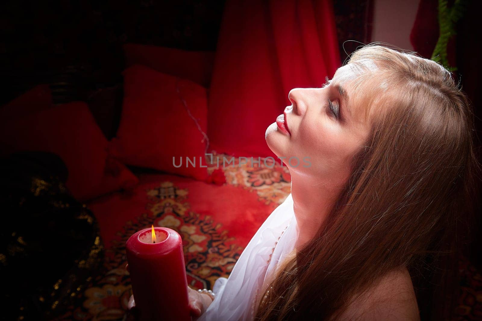 Beautiful arabian girl with candles in red room full of rich fabrics and carpets in sultan harem. Photo shoot of woman an oriental style odalisque. Model poses in sari as caring wife and hostess