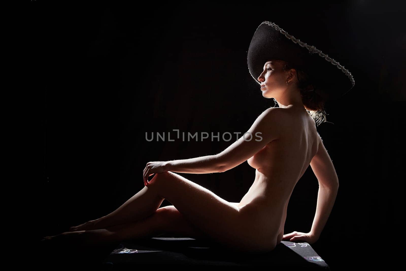 Silhouette of a beautiful young naced girl in a Mexican hat on a dark background. Slim exotic model posing naked in the studio. Body portrait of sexy girl in usial photoshoot by keleny