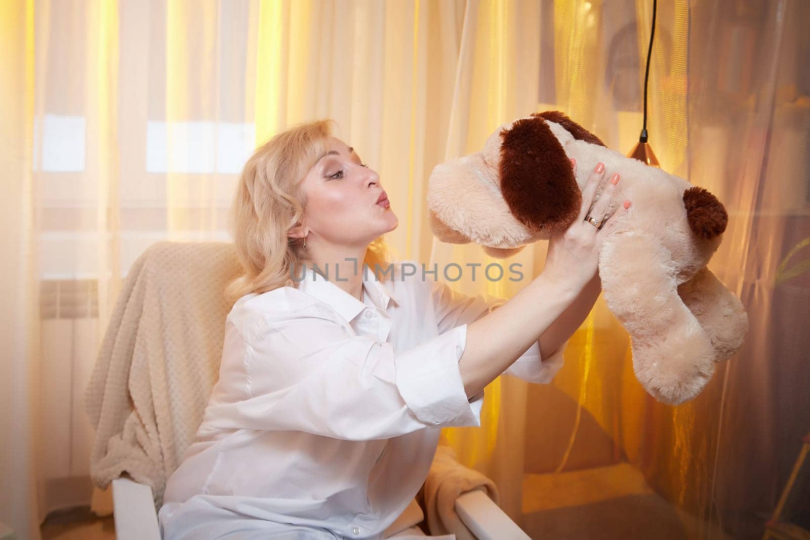 Adult mature woman of 40-60 years old with funny dog toy in a casual dress with white shirt and jeans. Room with calm cozy evening atmosphere with transparent curtains and soft warm light of lamps by keleny