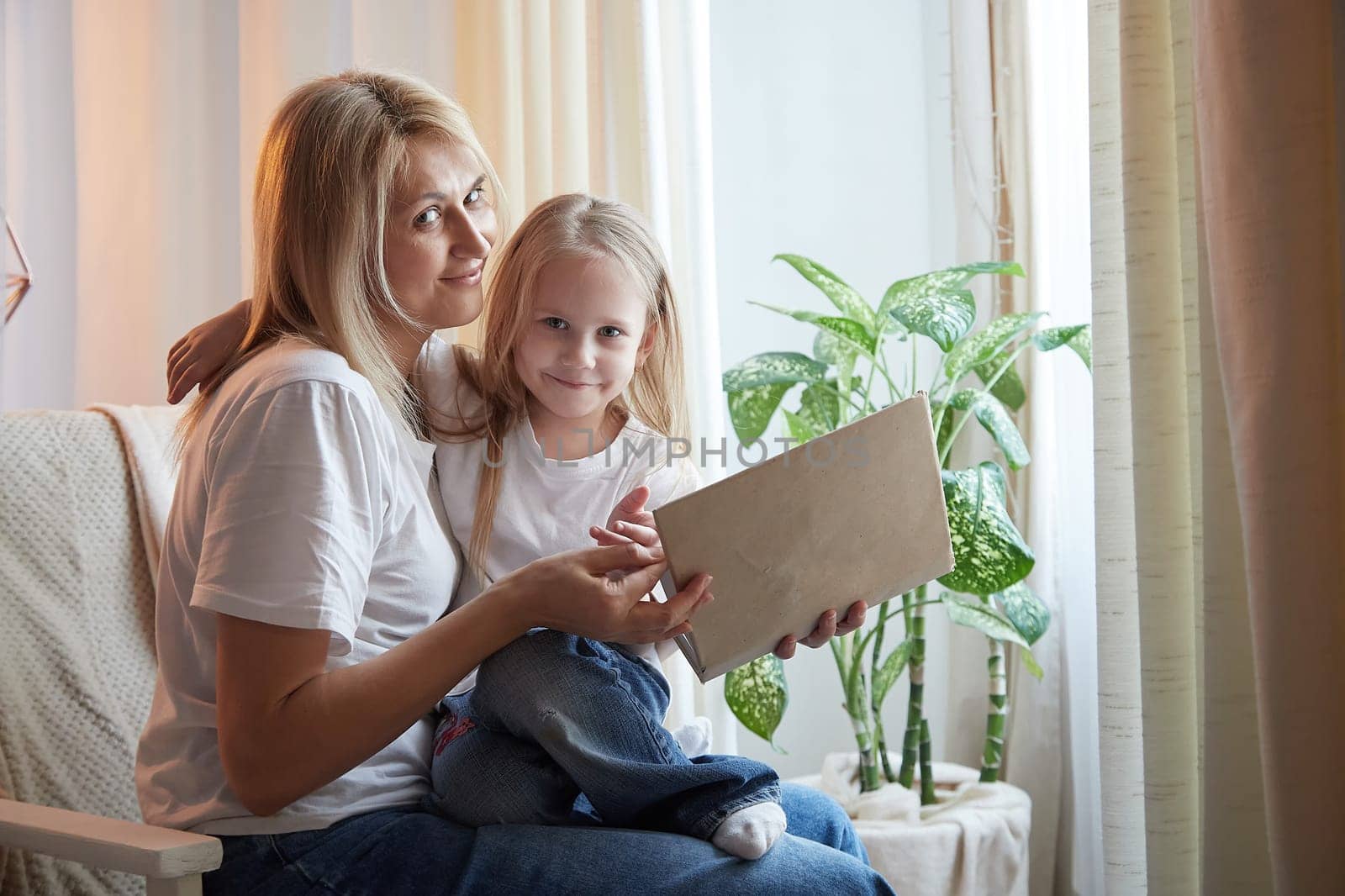 Happy loving family with mother and daughter reading book in living room. Woman mom and small child girl having convercation and studing inside of the home by keleny