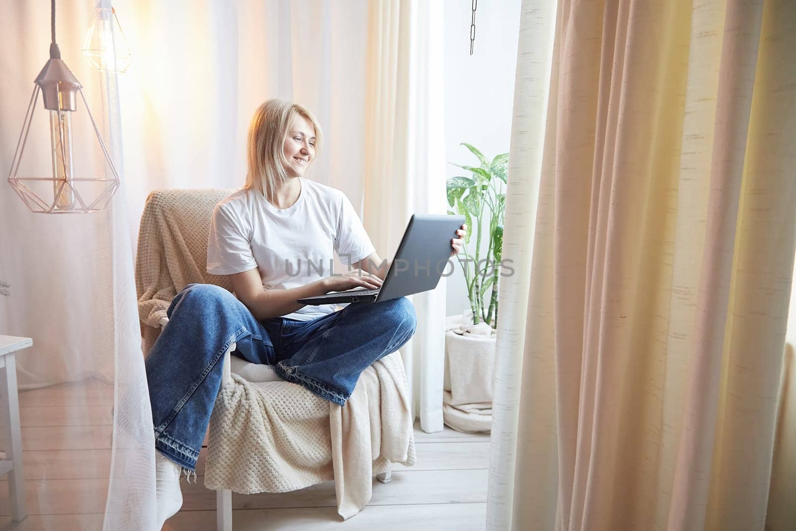 Young blonde woman using laptop computer at home. Girl is resting, chating and looking in camera near window at home. Nice atmosphere. The concept of cozy home