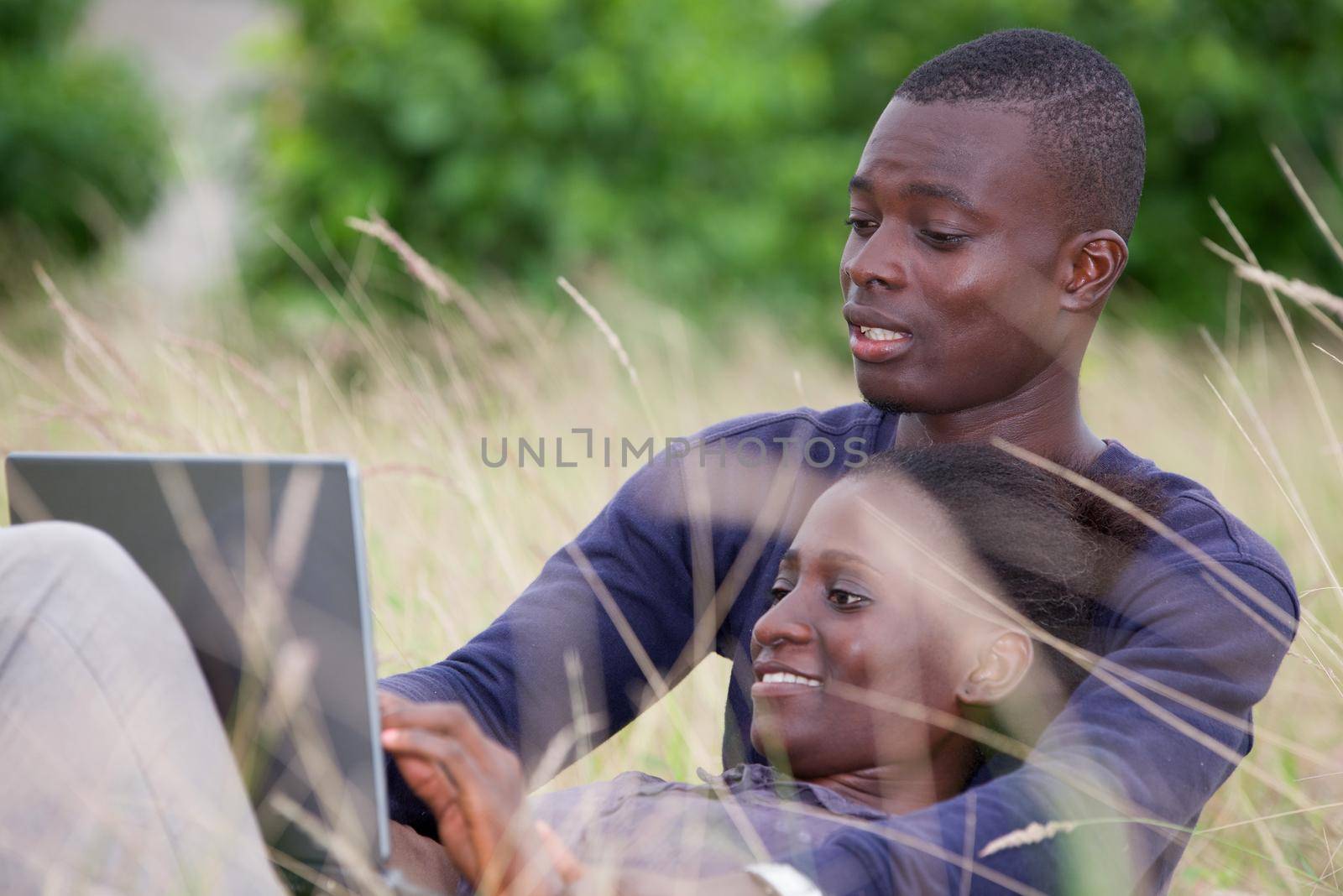 young african couple sitting in the grass with laptop and look at the screen smiling.