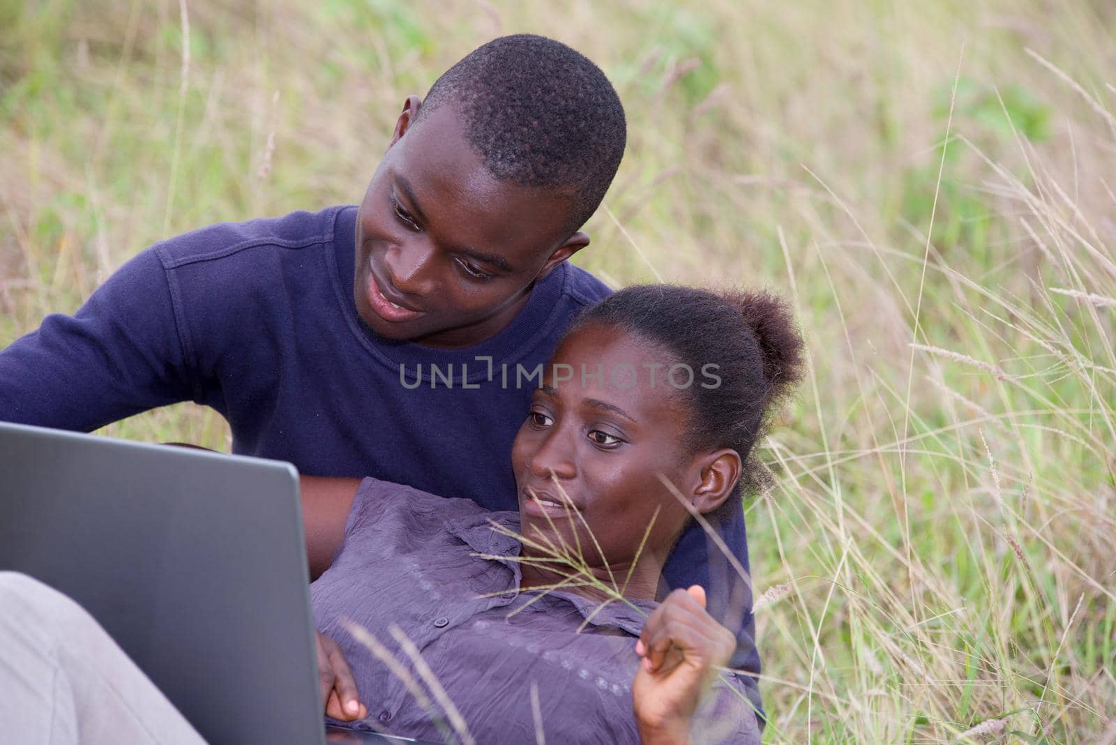 young african couple sitting in grass with laptop smiling.