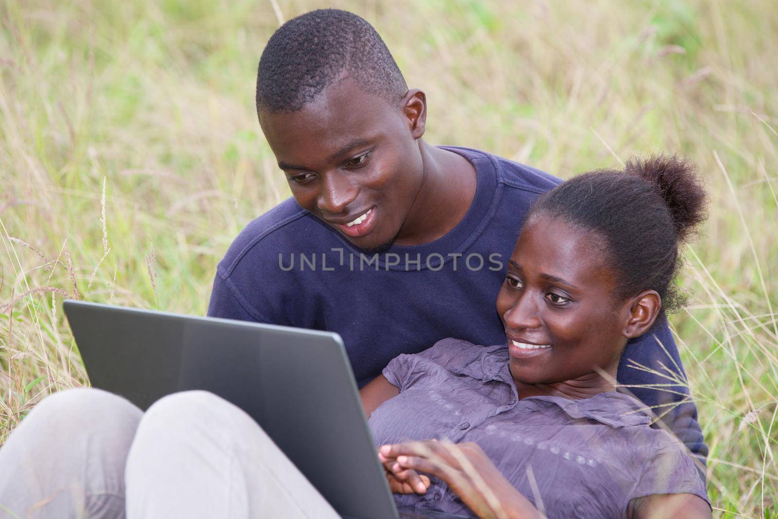 young african couple sitting in grass with laptop smiling.