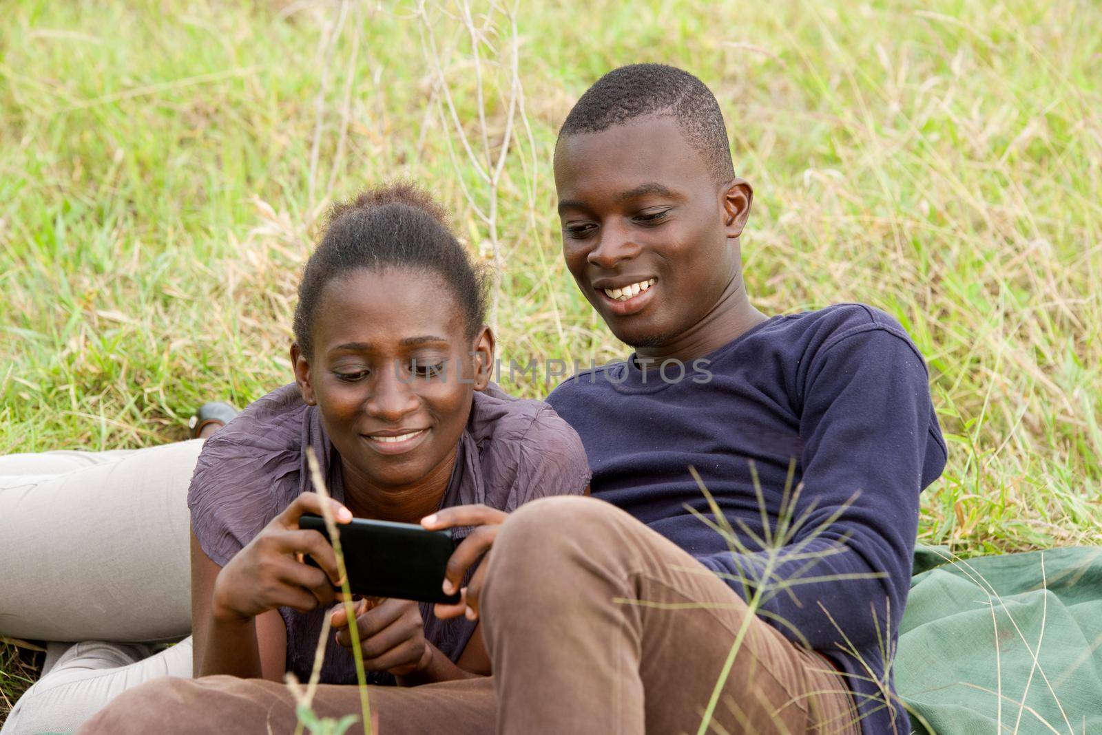 young african couple intertwined in a park looking at mobile phone while smiling.