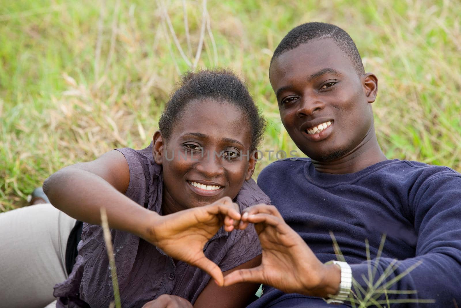 young african couple lying in grass making sign of hands while smiling at the camera.