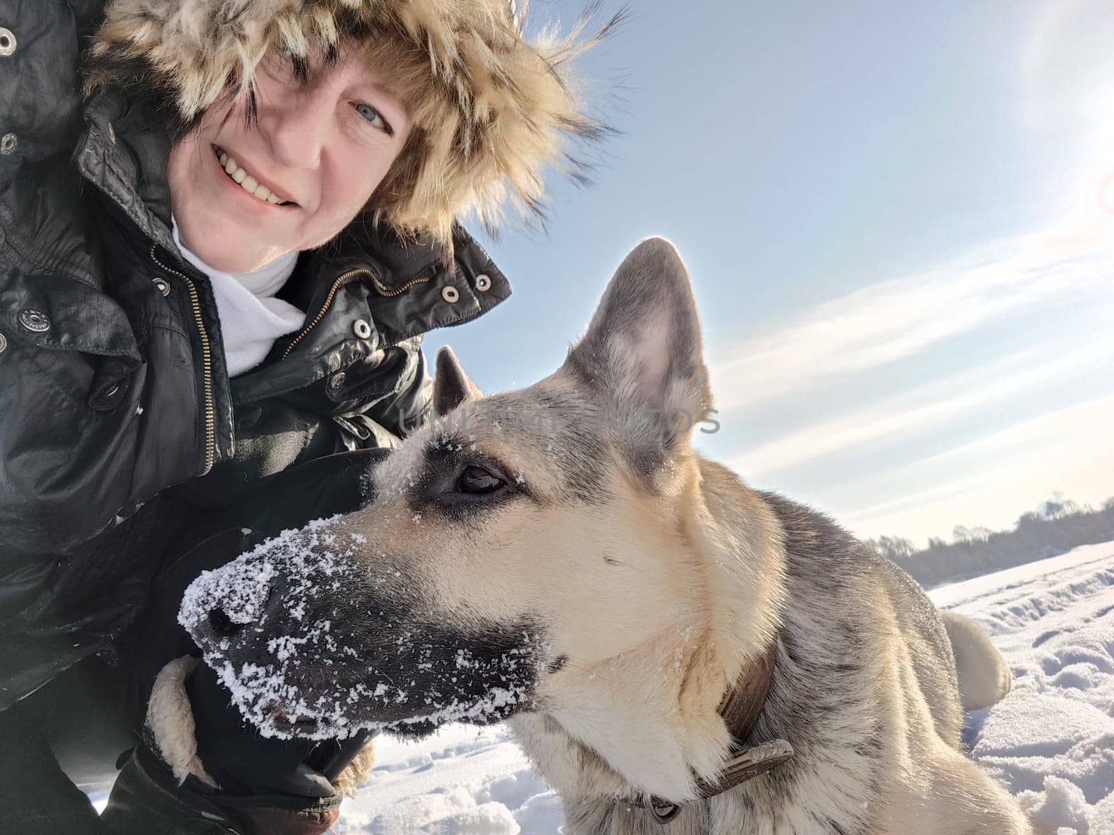 Adult girl or middle aged woman in a jacket with fluffy fur hood with shepherd dog in nature in winter on cold sunny day