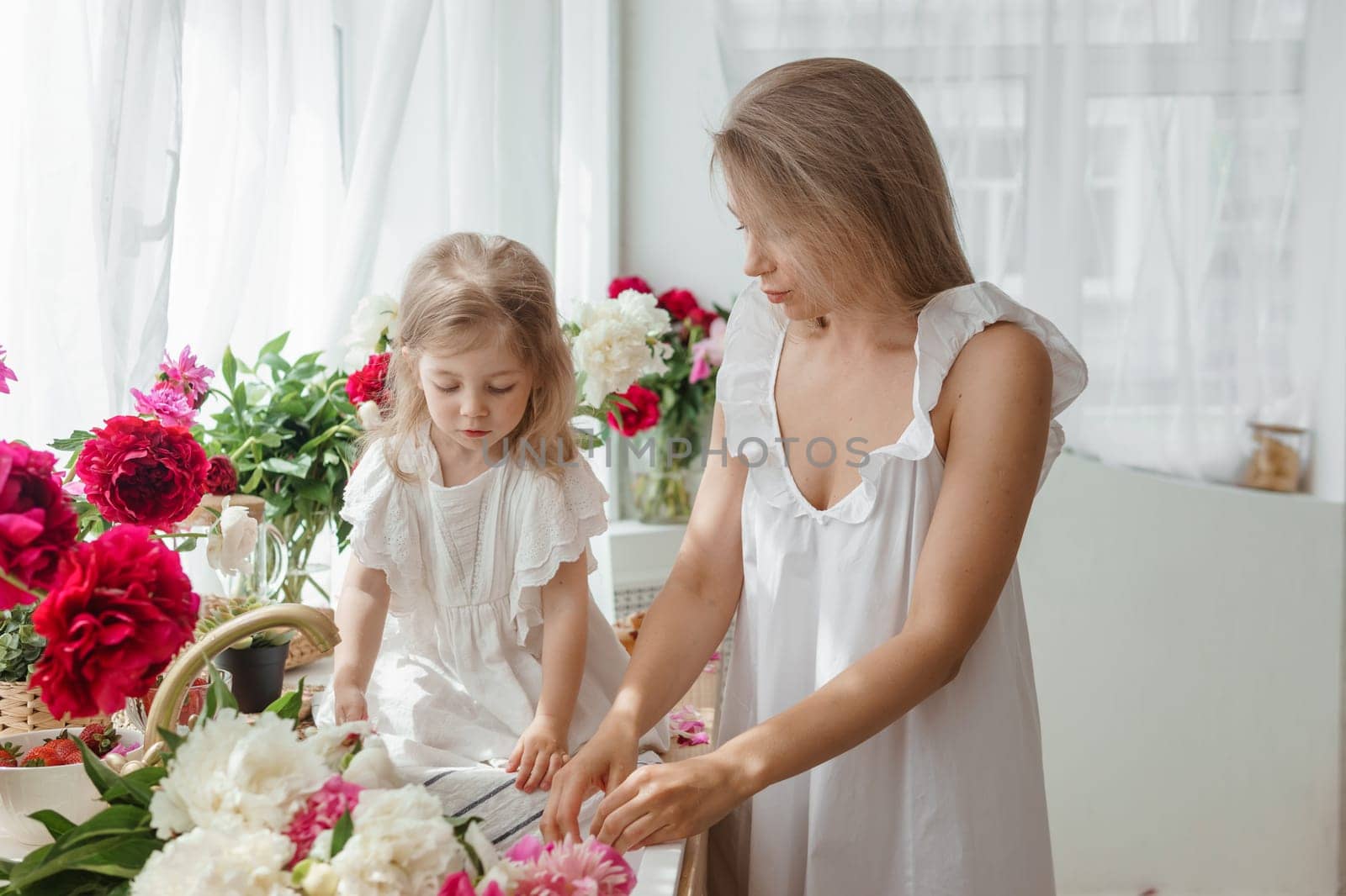 A little blonde girl with her mom on a kitchen countertop decorated with peonies. The concept of the relationship between mother and daughter. Spring atmosphere.