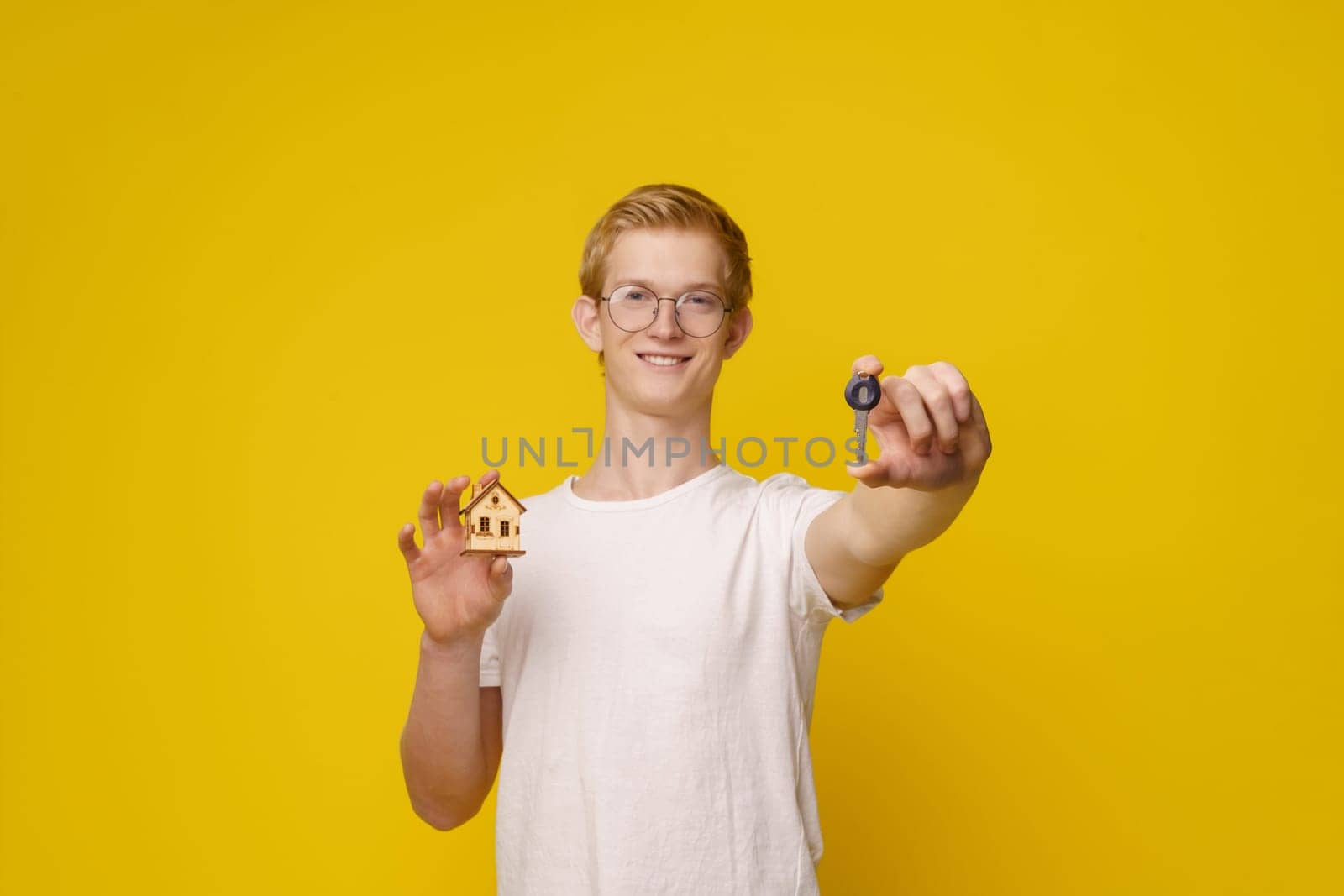 Young man stands against vibrant yellow background, holding set of keys and model house, symbolizing home ownership and the real estate industry. Focus on hand with key. by LipikStockMedia