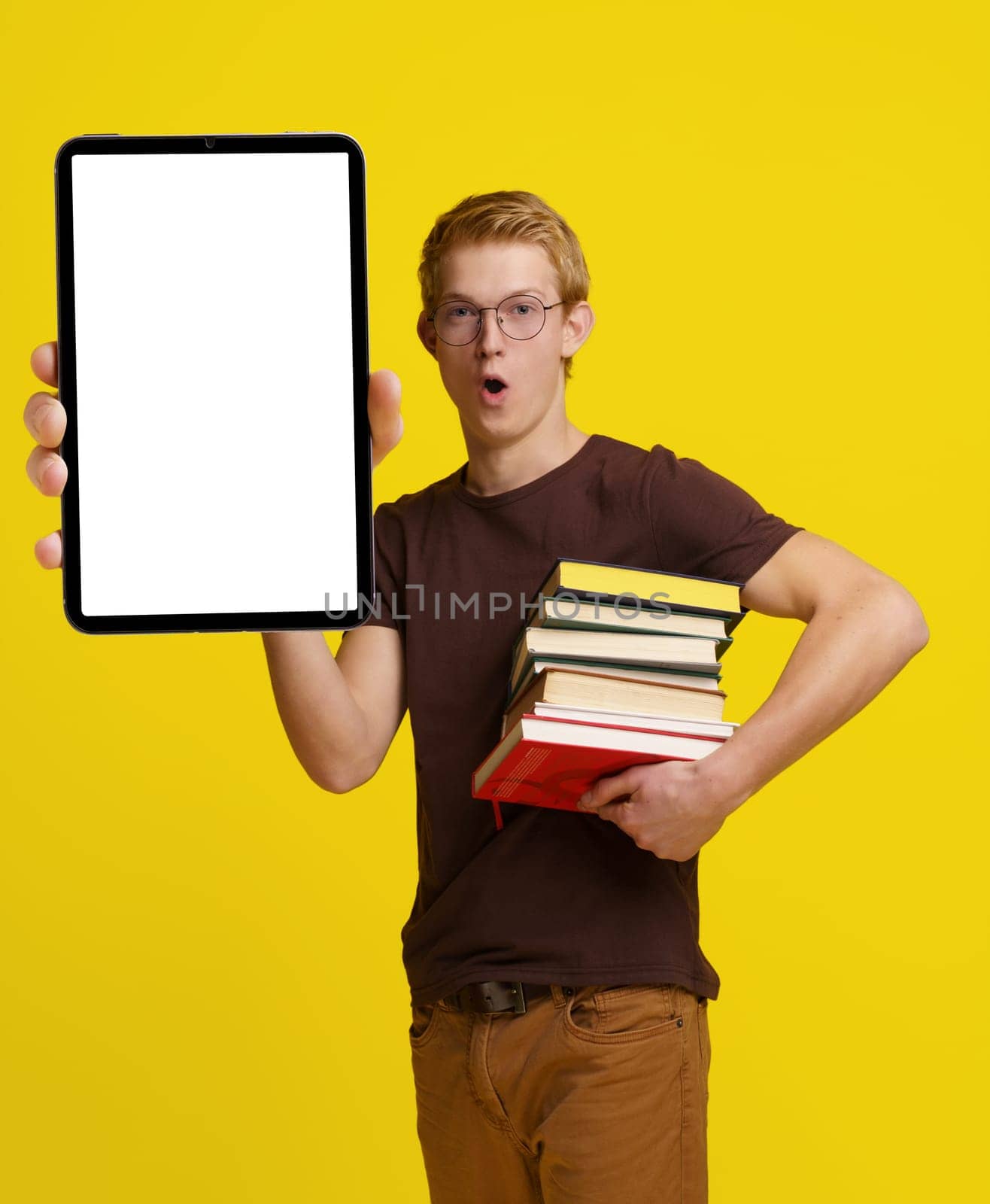Surprised student holding books and a Tablet PC with a blank white screen for copy space against a yellow background, showcasing the concept of modern education and the integration of technology in learning and studying. by LipikStockMedia