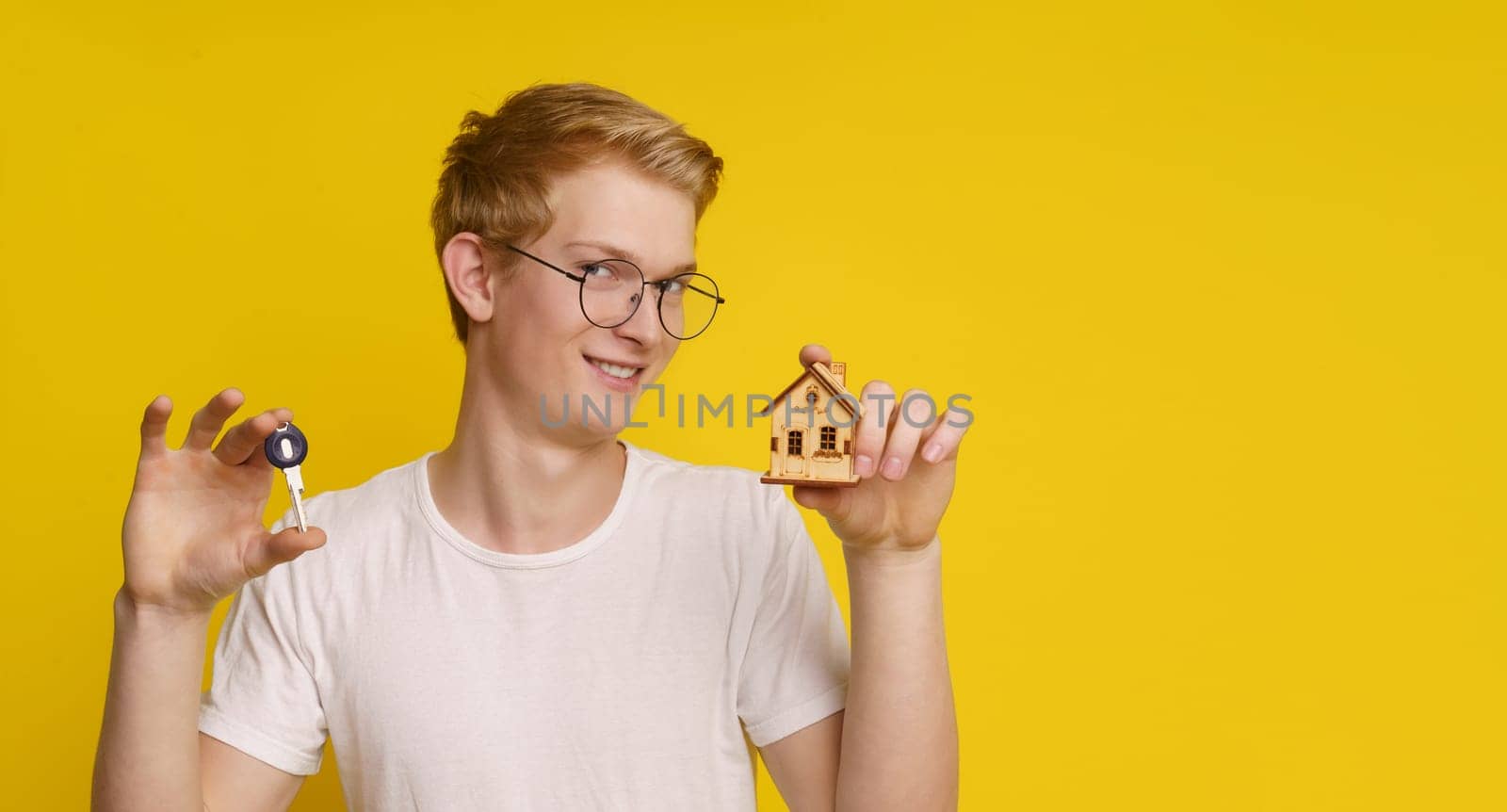 Young man holding a home architectural model, maquette and key against yellow background, showcasing the achievement of home ownership and success in the real estate market. by LipikStockMedia