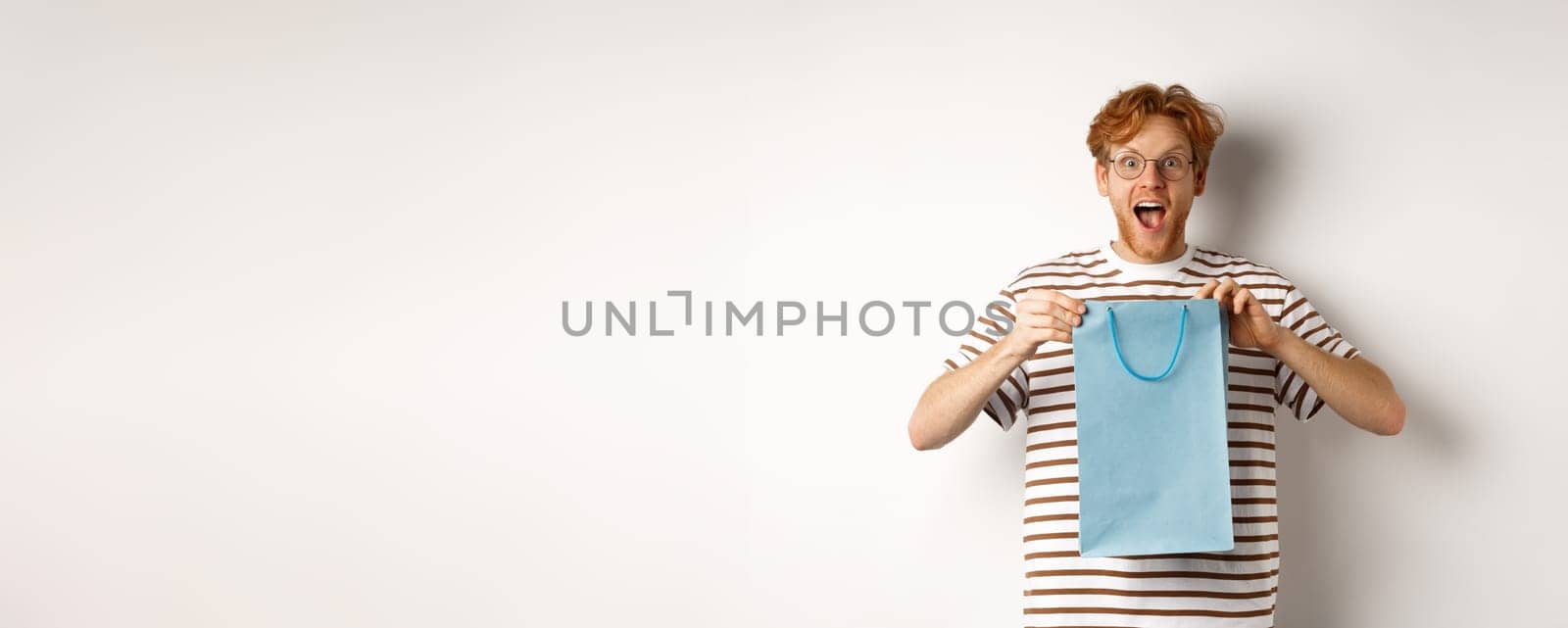 Holidays and celebration concept. Surprised redhead man receiving present inside shopping bag, looking amazed and thankful at camera, white background.