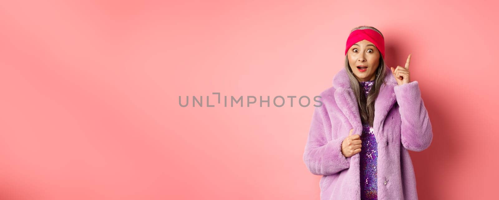Fashion and shopping concept. Smiling elderly woman having an idea. Stylish old lady in purple fake fur coat raising index finger, suggesting plan, standing excited on pink background.