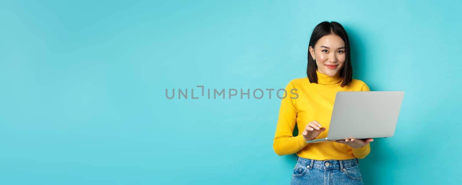 Young asian woman freelancer working on laptop and smiling, standing with computer over blue background.