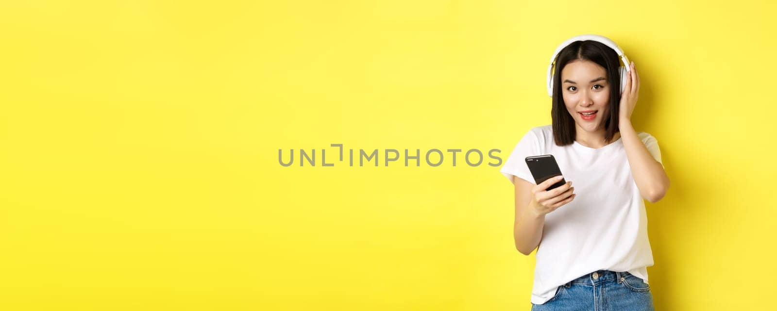 Modern asian woman listening music in wireless headphones, reading smartphone screen and smiling, standing in white t-shirt over yellow background.