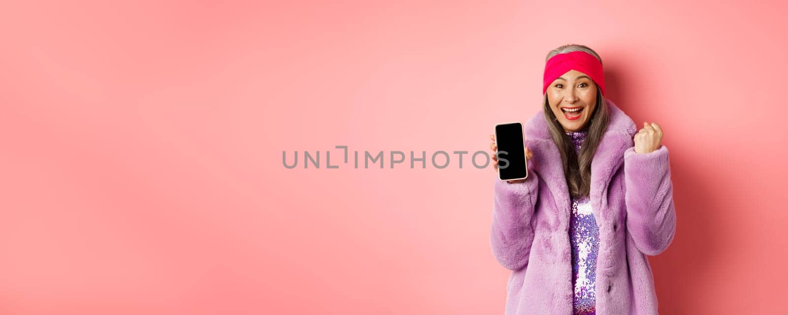 Online shopping and fashion concept. Happy asian senior woman winning prize in internet, showing blank mobile phone screen and making fist pump, scream of joy, standing over pink background.