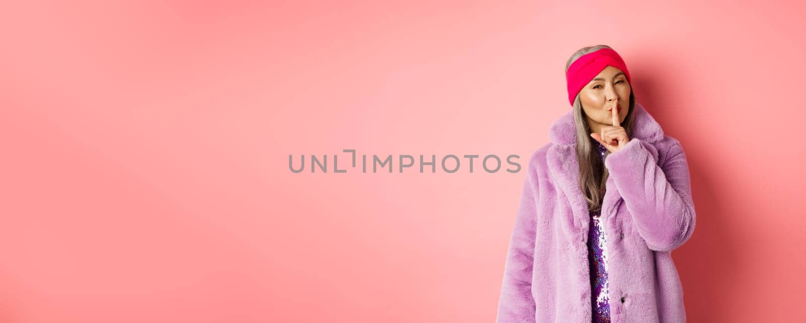 Fashion and shopping concept. Modern chinese senior woman shushing, telling a secret and looking cunning at camera, standing in trendy fake fur coat and headband on pink background.