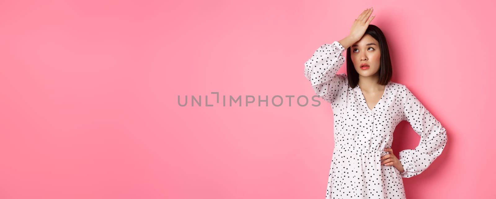 Annoyed and tired asian woman making facepalm, slap forehad and roll eyes disappointed, standing in white dress against pink background by Benzoix